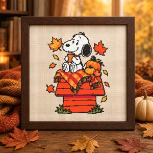 May include: A framed cross-stitch embroidery featuring a white beagle sitting on a red doghouse, holding an orange mug. The scene includes a plaid blanket, a pumpkin, and falling autumn leaves in shades of orange, yellow, and red on fabric.