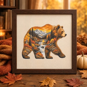 May include: A framed cross-stitch embroidery featuring a brown bear silhouette filled with a detailed autumn landscape. The scene inside the bear includes a flowing river, evergreen trees, golden foliage, and snow-capped mountains under a sunset sky.