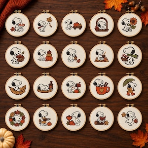 Puede incluir: Una colección de veinte bastidores de bordado circulares con diseños en punto de cruz de Snoopy en varias escenas temáticas de otoño, incluyendo calabazas, hojas caídas, girasoles y bebidas acogedoras sobre una superficie de madera.