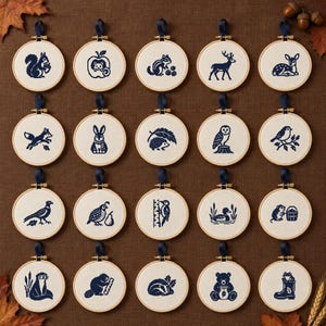 May include: A collection of twenty circular wooden embroidery hoops, each containing a cream-colored fabric panel with a dark blue cross-stitch design of a woodland animal or nature element, finished with a dark blue ribbon hanging loop.