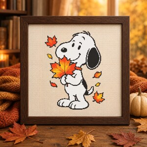 May include: A framed cross-stitch embroidery featuring a white cartoon dog with black ears holding a large, vibrant orange and yellow maple leaf. Several smaller autumn leaves in shades of red, orange, and yellow surround the dog on a cream fabric.