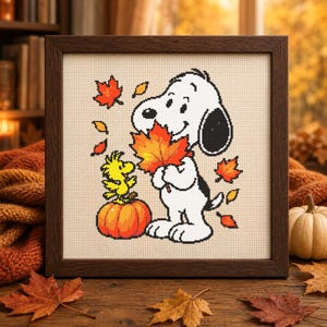 May include: A framed cross-stitch embroidery featuring a white beagle holding a large orange maple leaf, standing next to a small yellow bird perched on a round orange pumpkin, surrounded by scattered red and orange autumn leaves on a beige fabric.