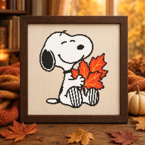 Könnte beinhalten: Ein gerahmtes Kreuzstich-Stickbild, das einen weißen Cartoon-Hund mit schwarzem Ohr und schwarzer Nase zeigt, der sitzt und ein Bündel leuchtend orangefarbener und roter Herbst-Ahornblätter vor einem hellbeigen Stoffhintergrund hält.