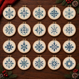 May include: Twenty circular wooden embroidery hoops containing cross-stitch patterns of blue and teal snowflakes on white fabric. Each hoop features a unique geometric snowflake design, arranged in a grid of four rows and five columns.