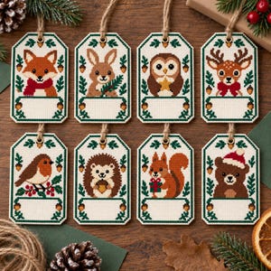 Op de afbeelding: Acht rechthoekige kruissteek cadeaulabels met bosdieren zoals een vos, konijn, uil, hert, roodborstje, egel, eekhoorn en beer. Elk label heeft een wit vlak om op te schrijven, groene bladranden en een ophanglus van jute touw.