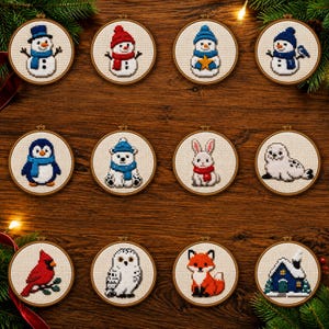 May include: Twelve circular wooden embroidery hoops containing cross-stitch patterns of winter themes. Designs include snowmen, a penguin, a polar bear, a rabbit, a seal, a cardinal, an owl, a fox, and a snow-covered house on white fabric.