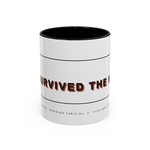 May include: A white ceramic mug with a black interior and rim. The exterior features horizontal black lines and the text "SURVIVED THE M" in bold, dark orange letters with a black shadow effect. Smaller text reads "WEDGING TABLE No. 3".