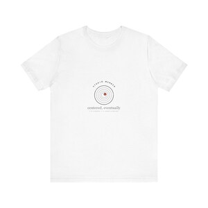 May include: A white short-sleeve t-shirt featuring a graphic on the chest. The graphic displays concentric black circles with a small red dot, the text STUDIO MEMBER arched above, and the text centered, eventually below the circles.