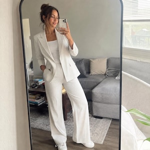 May include: A white blazer and matching wide-leg trousers featuring thin vertical pinstripes, paired with a plain white scoop-neck tank top and white trainers. The blazer has a notched lapel, long sleeves, and two front pockets.