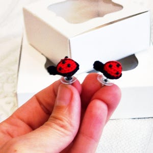 Puede incluir: Un par de pendientes de botón con diseños de pequeñas mariquitas de ganchillo. Cada mariquita tiene un cuerpo rojo brillante con puntos negros y una cabeza negra, unida a un poste de plástico transparente para orejas perforadas.