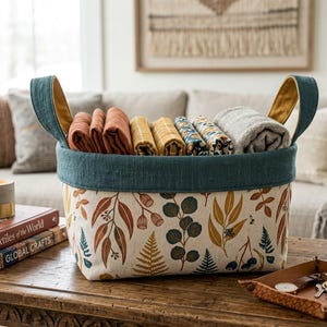 May include: A fabric storage basket with a cream-coloured exterior featuring a botanical print of leaves and ferns in shades of rust, mustard yellow, and dark teal. The basket has a solid dark teal rim and two matching fabric handles.
