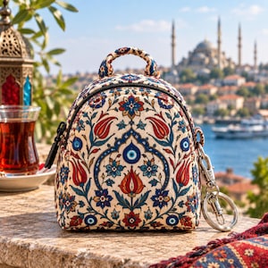 May include: A small backpack featuring a cream-colored base with a repeating floral pattern in red, blue, and green. The design includes stylized tulip motifs and circular blue and white evil eye symbols. It has a top handle and a silver metal keyring.