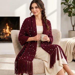 May include: A deep burgundy knitted shawl featuring a ribbed texture, scattered white pearl-like embellishments, and long fringe trim along the bottom edges. The garment is draped over the shoulders and hangs down the front.