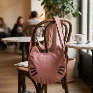 Pode incluir: Uma bolsa de ombro de lona em tom rosa seco com formato de cabeça de gato, apresentando duas orelhas pontiagudas no topo e três bigodes horizontais pretos bordados em cada lado da parte inferior frontal da bolsa.