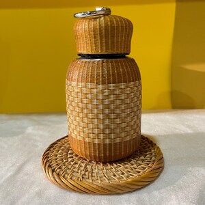 Puede incluir: Un recipiente cilíndrico con un exterior de ratán tejido en tonos marrón y crema y una tapa a juego con un asa de metal plateado. El recipiente descansa sobre un posavasos circular de ratán tejido con un borde grueso y trenzado.