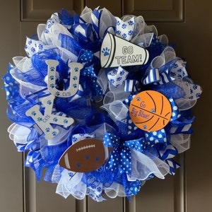 May include: A blue and white wreath with a basketball, football, and megaphone. The wreath is decorated with blue and white ribbons and has the letters "UK" and the word "CATS" on it. The megaphone says "GO TEAM!" and the basketball says "Go Big Blue!"
