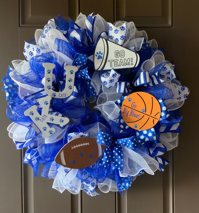 May include: A blue and white wreath with a basketball, football, and megaphone. The wreath is decorated with blue and white ribbons and has the letters "UK" and the word "CATS" on it. The megaphone says "GO TEAM!" and the basketball says "Go Big Blue!"
