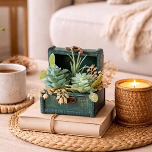 May include: A teal wooden treasure chest planter with faux succulents and greenery, sitting atop a stack of books. A lit candle in a woven holder and a mug of coffee are also on the table, creating a cozy, decorative scene.