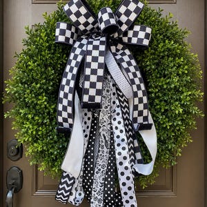 May include: A decorative wreath featuring a lush green base adorned with a large bow. The bow is made of black and white checkered ribbon, with additional ribbons in polka dot, floral, and chevron patterns. The wreath is displayed on a brown door.