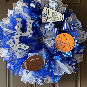 May include: A blue and white wreath with the letters "UK" and the words "Go Big Blue!" and "Go Team!" on it. The wreath is decorated with blue and white ribbons, white mesh, and paw prints.