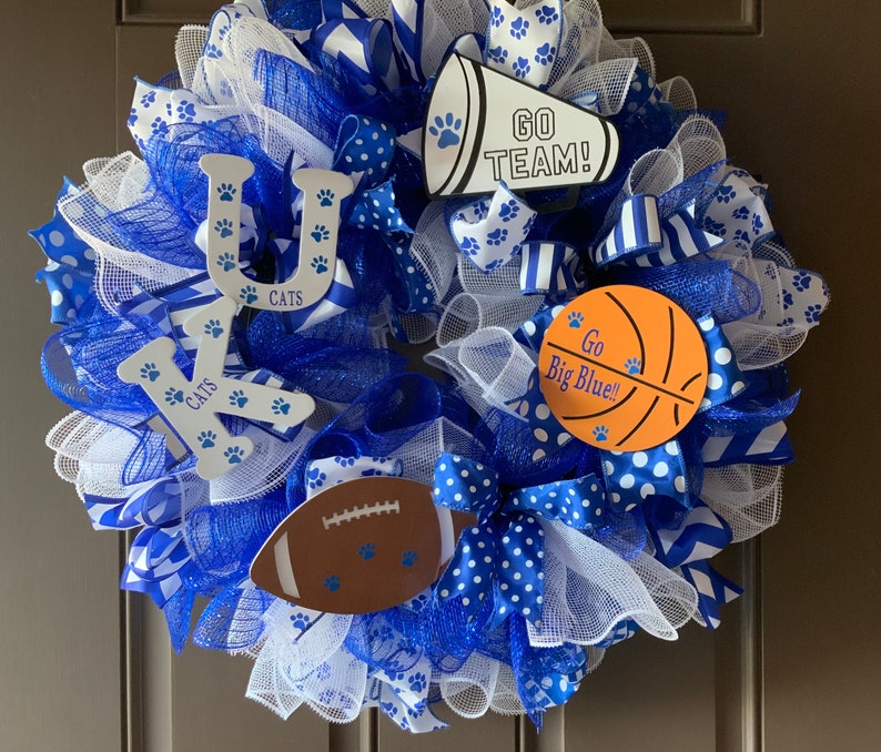 May include: A blue and white wreath with the letters "UK" and the words "Go Big Blue!" and "Go Team!" on it. The wreath is decorated with blue and white ribbons, white mesh, and paw prints.