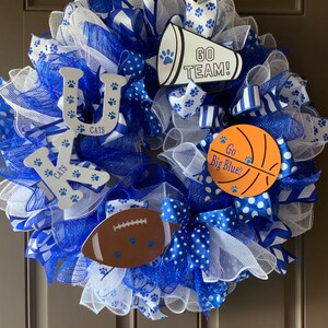 May include: A blue and white wreath with the letters "UK" and the words "Go Big Blue!" and "Go Team!" on it. The wreath is decorated with blue and white ribbons, white mesh, and paw prints.