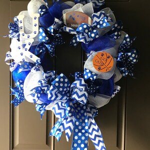May include: A blue and white wreath with a football, basketball, and paw print decorations. The wreath has a blue and white ribbon with white polka dots and a chevron pattern. The wreath also has a sign that says "Go Cats!"