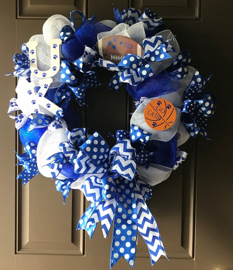 May include: A blue and white wreath with a football, basketball, and paw print decorations. The wreath has a blue and white ribbon with white polka dots and a chevron pattern. The wreath also has a sign that says "Go Cats!"