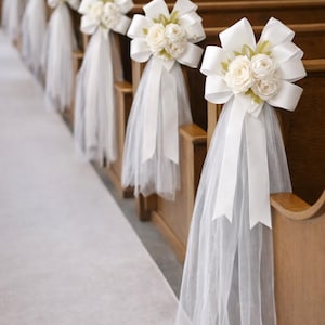 May include: White floral pew decorations with large bows and tulle. The decorations are attached to wooden pews in a church. The aisle is covered with a white carpet.
