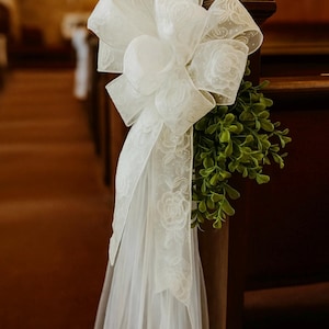 May include: A large white bow with floral lace detailing and a long, sheer tulle drape. The bow is attached to a wooden pew and accented with green foliage, creating a classic wedding decoration.