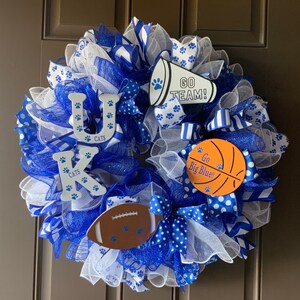 May include: A blue and white wreath with the letters "UK" and the word "CATS" on it. The wreath also has a blue and white megaphone with the words "GO TEAM!" on it, a basketball with the words "Go Big Blue!" on it, and a football with paw prints on it.