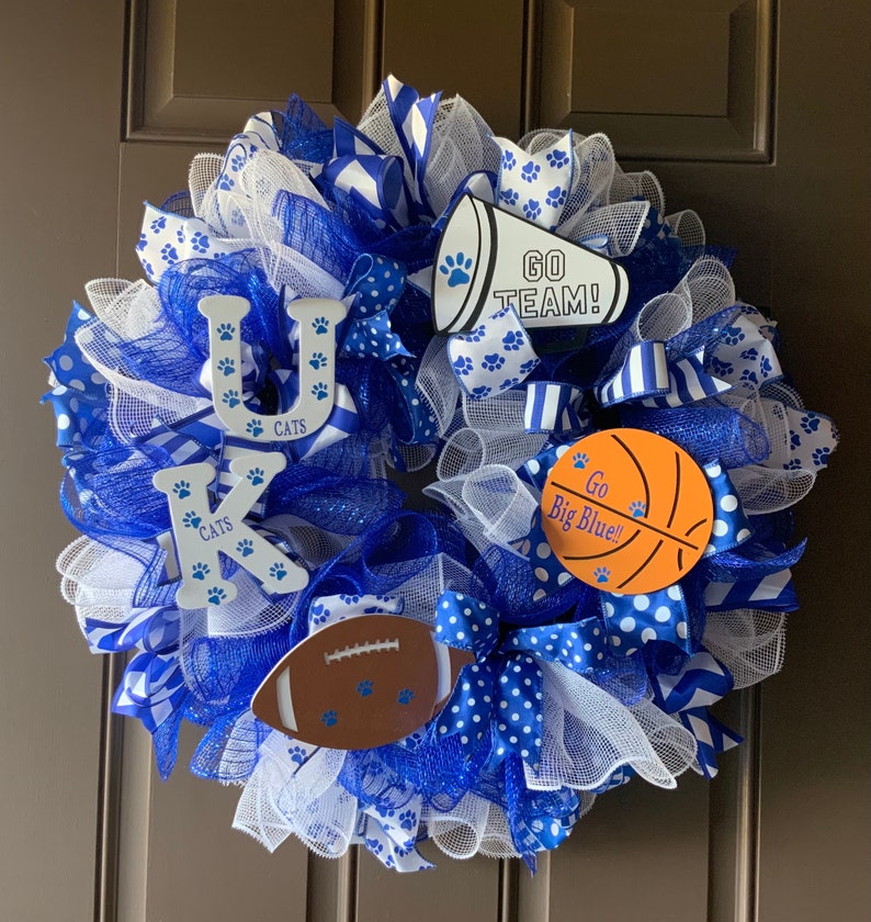 May include: A blue and white wreath with the letters "UK" and the word "CATS" on it. The wreath also has a blue and white megaphone with the words "GO TEAM!" on it, a basketball with the words "Go Big Blue!" on it, and a football with paw prints on it.