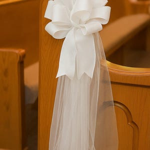 May include: A white satin bow with long, flowing tulle is attached to a wooden pew. The bow is large and decorative, with multiple loops and tails. The tulle hangs down, adding a touch of elegance to the wedding decor.