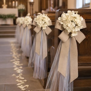 May include: Wedding ceremony decorations featuring white floral arrangements with burlap bows and sheer fabric accents attached to wooden pews. The aisle is adorned with scattered white flower petals.