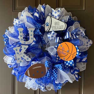 May include: A blue and white wreath with the letters UK, a basketball, a football, and a megaphone with the text "Go Team!" and "Go Big Blue!" The wreath is decorated with blue and white ribbons and mesh.