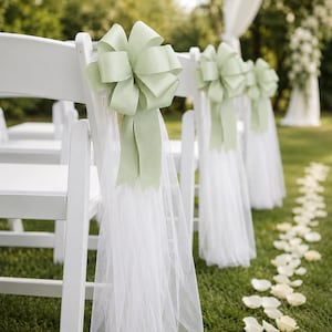 May include: A row of white folding chairs decorated with sage green satin bows and sheer white tulle fabric drapes. Each chair features a large, multi-looped ribbon tied to the backrest, with the tulle extending downward toward the grass.