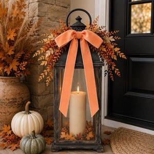 May include: A black metal lantern featuring a large, light orange velvet bow at the top, accented with artificial autumn foliage and berries. Inside the lantern, a single cream-colored pillar candle is lit, surrounded by small orange maple leaves.