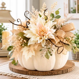 May include: A decorative arrangement featuring a large white pumpkin base topped with cream-colored dahlia flowers, beige ribbon, green leaves, small white berries, and dark brown curly twigs. The floral display is centered on a round wooden base.