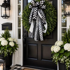 May include: A black front door with a green wreath and a large black and white checkered bow. The door is flanked by black lanterns, planters with white flowers, and a welcome mat with the word "WELCOME".
