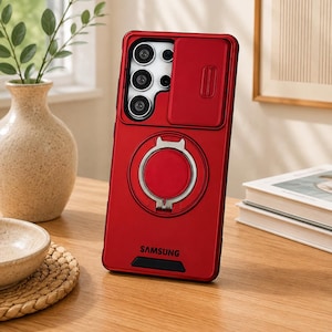 May include: A red smartphone case for a Samsung device featuring a sliding camera lens cover, a circular metal ring stand on the back, and the word SAMSUNG printed in black at the bottom. The case has a matte finish with black accents.