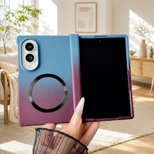Op de afbeelding: Een opvouwbaar smartphonehoesje met een blauw-naar-paarse gradiëntafwerking. Het achterpaneel bevat een witte camerabehuizing met twee ronde lenzen en een zwarte magnetische ring. Het hoesje is geopend en toont een zwart scherm aan de binnenkant.