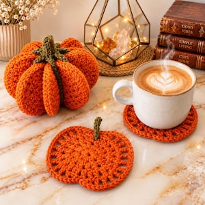 May include: A crocheted orange pumpkin with a green stem, a matching flat crocheted pumpkin coaster, and a white ceramic mug filled with coffee featuring latte art. The mug rests on a second orange crocheted pumpkin coaster.