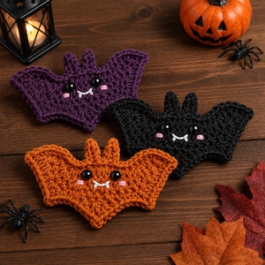 May include: Three crocheted bat ornaments in purple, black, and orange. Each bat features two black plastic eyes, embroidered pink cheeks, and two small white fangs. The bats have pointed ears and wide, textured wings, crafted with a consistent yarn stitch pattern.