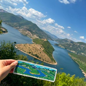 Puede incluir: Un marcapáginas rectangular de punto de cruz con un diseño de paisaje detallado que muestra un río azul serpenteante, colinas verdes y un borde de ganchillo blanco. El bordado representa una vista de valle con hilos azules y verdes.