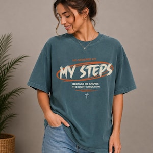 May include: A dark teal short-sleeve t-shirt featuring the text HE ORDERED MY MY STEPS BECAUSE HE KNOWS THE RIGHT DIRECTION in white and orange, with a small white cross graphic centered below the text on the front of the garment.