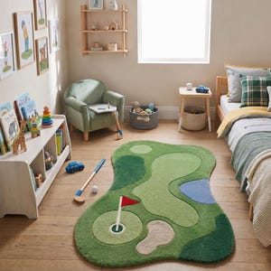 Peut inclure: Un tapis vert en forme de terrain de golf, avec des nuances de vert, un bunker de sable beige, un obstacle d'eau bleu et un trou de golf blanc avec un drapeau rouge. Le tapis est posé sur un sol en bois dans une pièce avec un lit et une chaise.