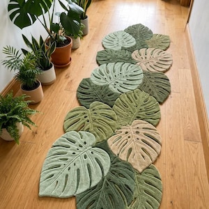 May include: A runner rug shaped like a collection of overlapping monstera leaves in various shades of green, sage, and beige. The rug features detailed cutouts and textured veins mimicking the appearance of tropical foliage on a floor.