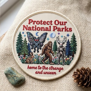 May include: A round embroidered patch with a cream background featuring the red text Protect Our National Parks and home to the strange and unseen. It depicts a moth, a rabbit, a sasquatch, and a deer-headed creature among pine trees.