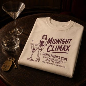 May include: A cream-colored t-shirt featuring a dark brown graphic of a pin-up figure next to a martini glass. The text reads MIDNIGHT CLIMAX GENTLEMEN'S CLUB, DON'T MIND THE FUZZ - FEEL THE BUZZ, 225C CHESTNUT ST. SAN FRANCISCO in a retro font.