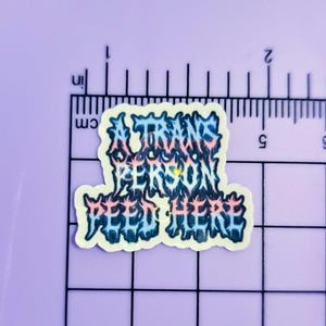 May include: A die-cut sticker featuring the text A TRANS PERSON PEED HERE in a jagged, metal-style font with a blue, pink, and white gradient. The sticker is positioned against a black grid ruler, showing a width of approximately 2.25 inches.